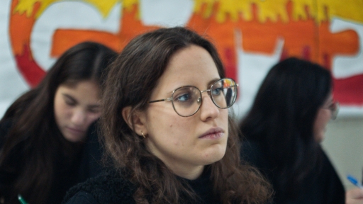 The image shows a young woman wearing glasses, her expression focused and serious, as if paying close attention. She’s surrounded by others in the background, similarly engaged in their activity, in an environment that suggests a meeting or workshop setting.