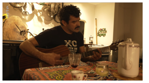 On voit un homme chez lui, guitare à la main.