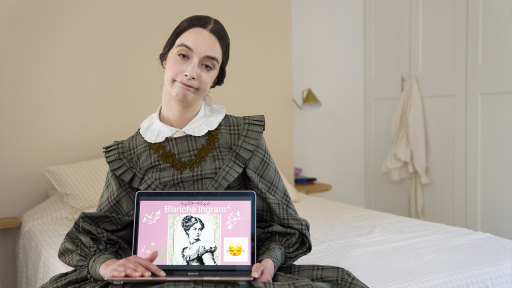 On voit la comédienne déguisée en Jane Eyre, entrain de faire une présentation sur son ordinateur. Elle est assise sur son lit. 