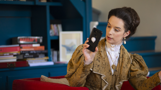 On voit la comédienne déguisée en Nora Helmer, dans son salon, qui regarde quelque chose sur son téléphone.