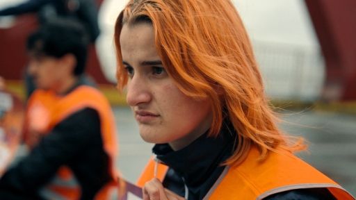 The image shows a person with vivid orange hair wearing a high-visibility safety vest, their expression intense and focused. They’re looking off into the distance, with a blurred urban background behind them. The scene suggests they’re part of a blockade at a protest.