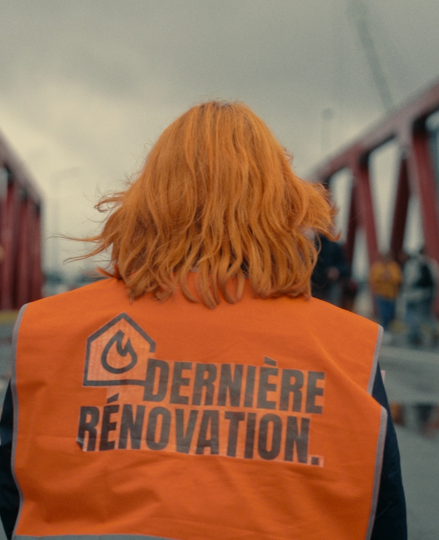 L'image montre une personne vue de dos, avec des cheveux orange vif, portant un gilet de sécurité orange sur lequel est inscrit "DERNIÈRE RÉNOVATION" accompagné d'un symbole de flamme. Elle se tient sur un pont rouge, où plusieurs autres personnes sont visibles au loin, floues, sous un ciel gris. Le sol est humide, comme après la pluie, reflétant les structures et les silhouettes. L’atmosphère suggère un contexte de mobilisation ou de manifestation.