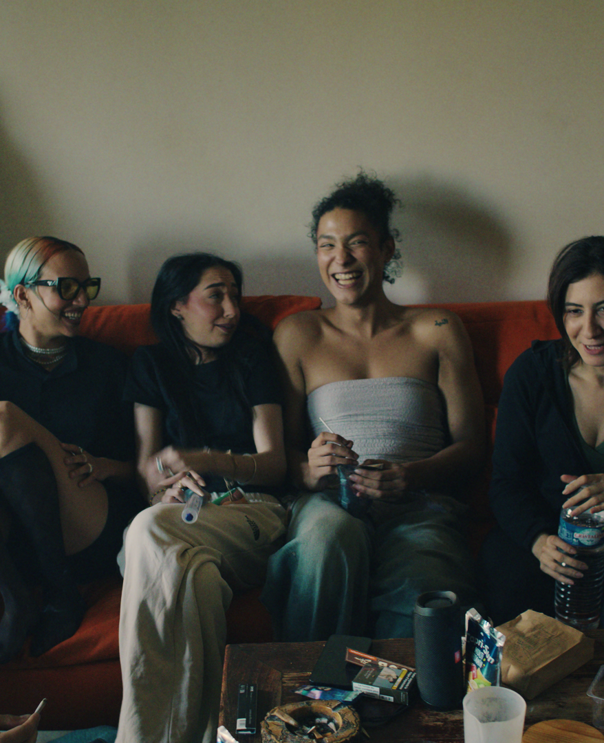 L'image montre un groupe de jeunes assis ensemble sur un canapé dans un salon, partageant un moment de convivialité et de détente. Ils rient et discutent, chacun affichant des expressions détendues et joyeuses. Certains tiennent des boissons ou des cigarettes.